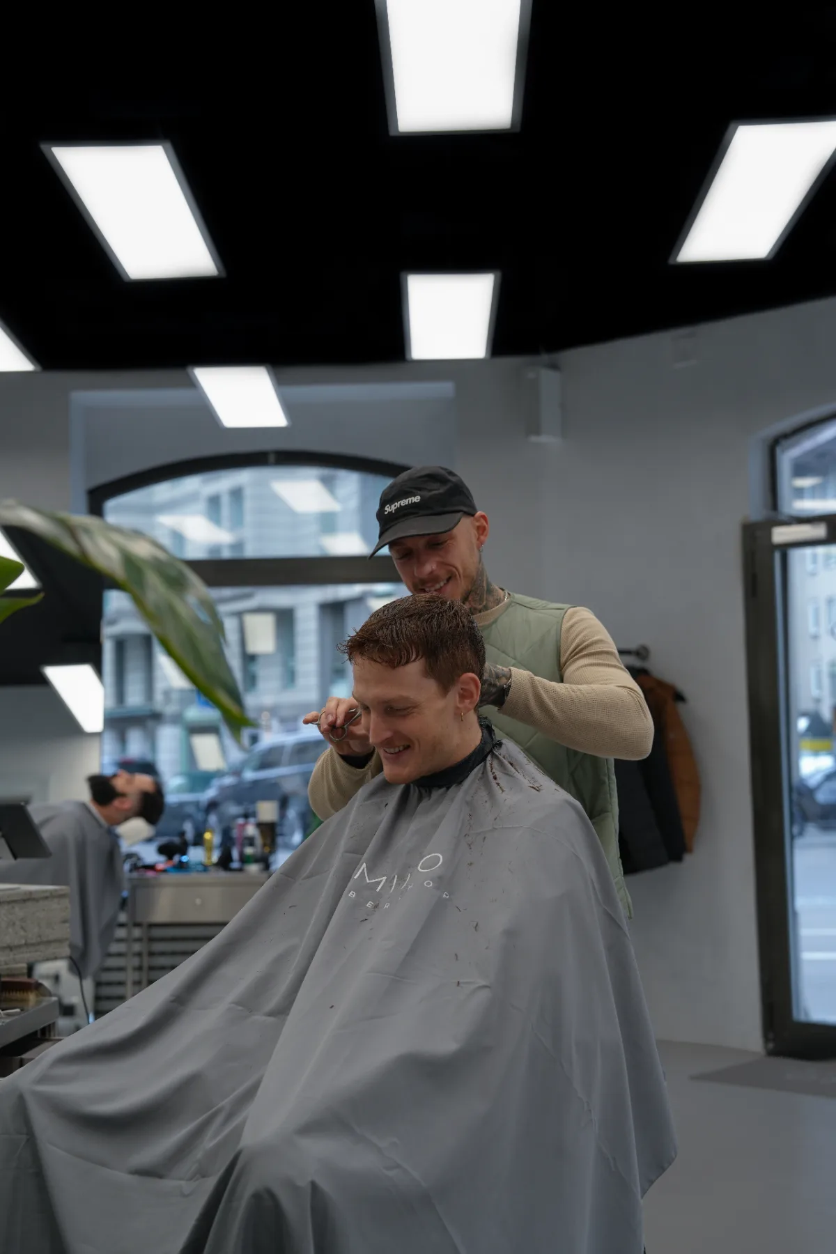 Brand photograph of a barber trimming a client's hair with scissors in a bright barbershop interior.