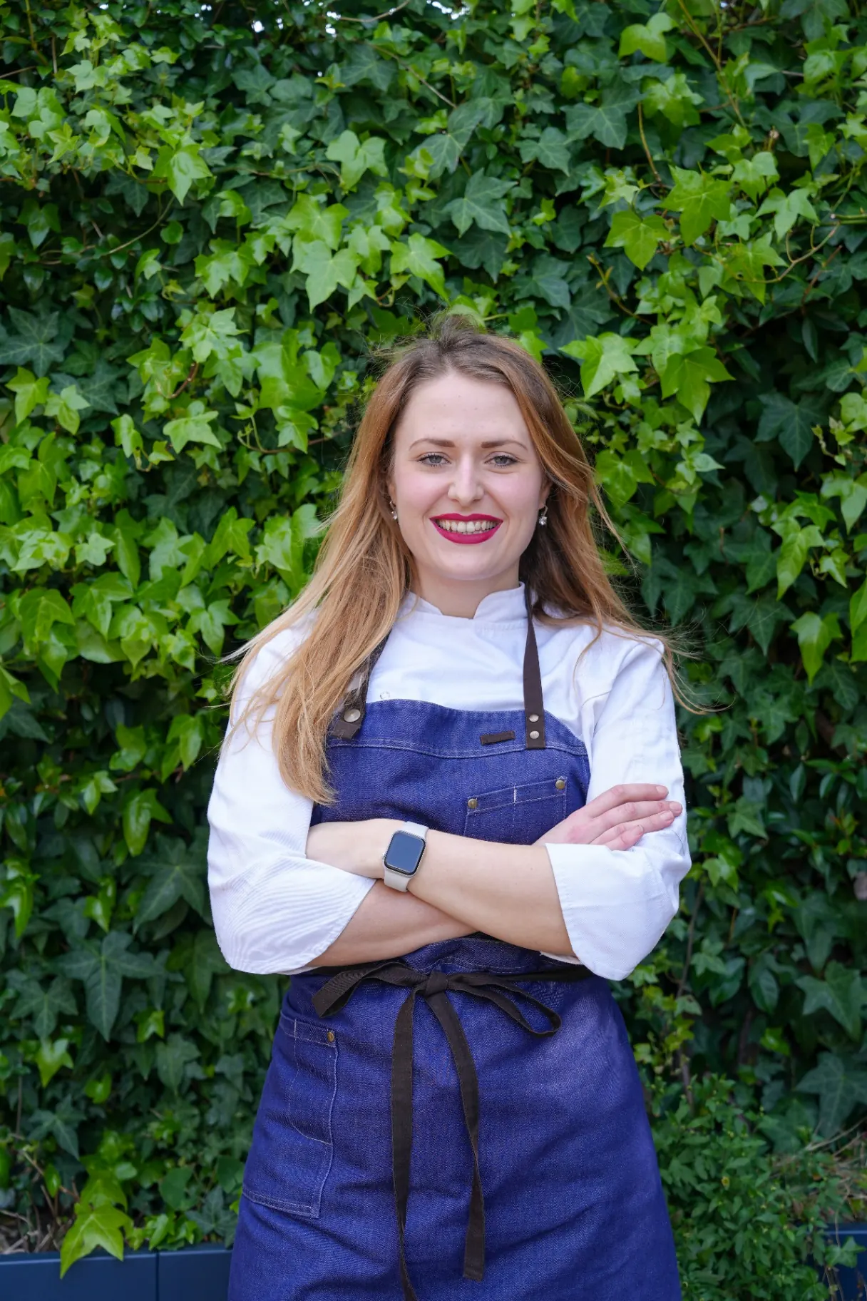 Editorial portrait of a chef in a denim apron standing against a lush ivy wall.