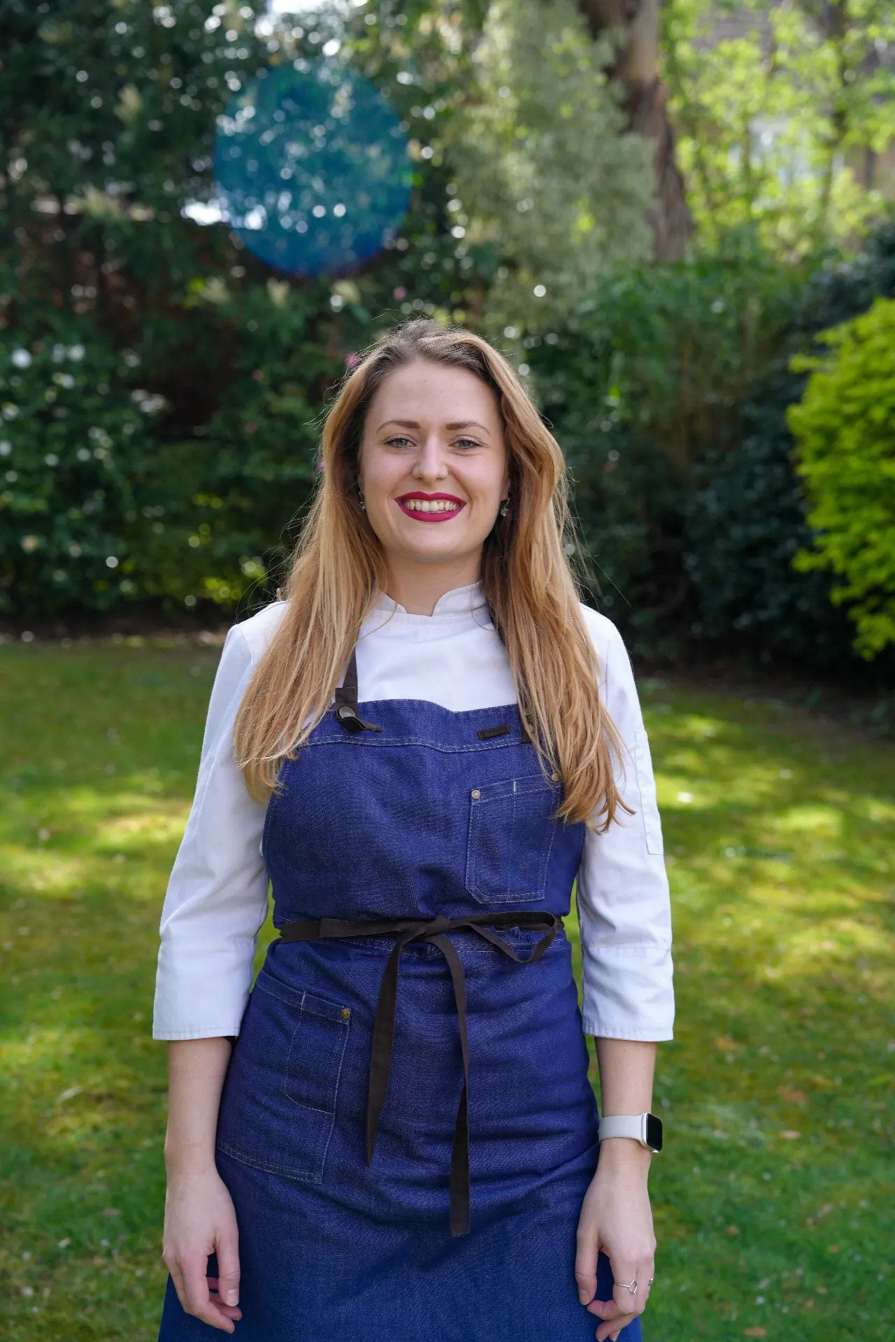 Full-length portrait of a chef in a blue apron standing outdoors in natural light.
