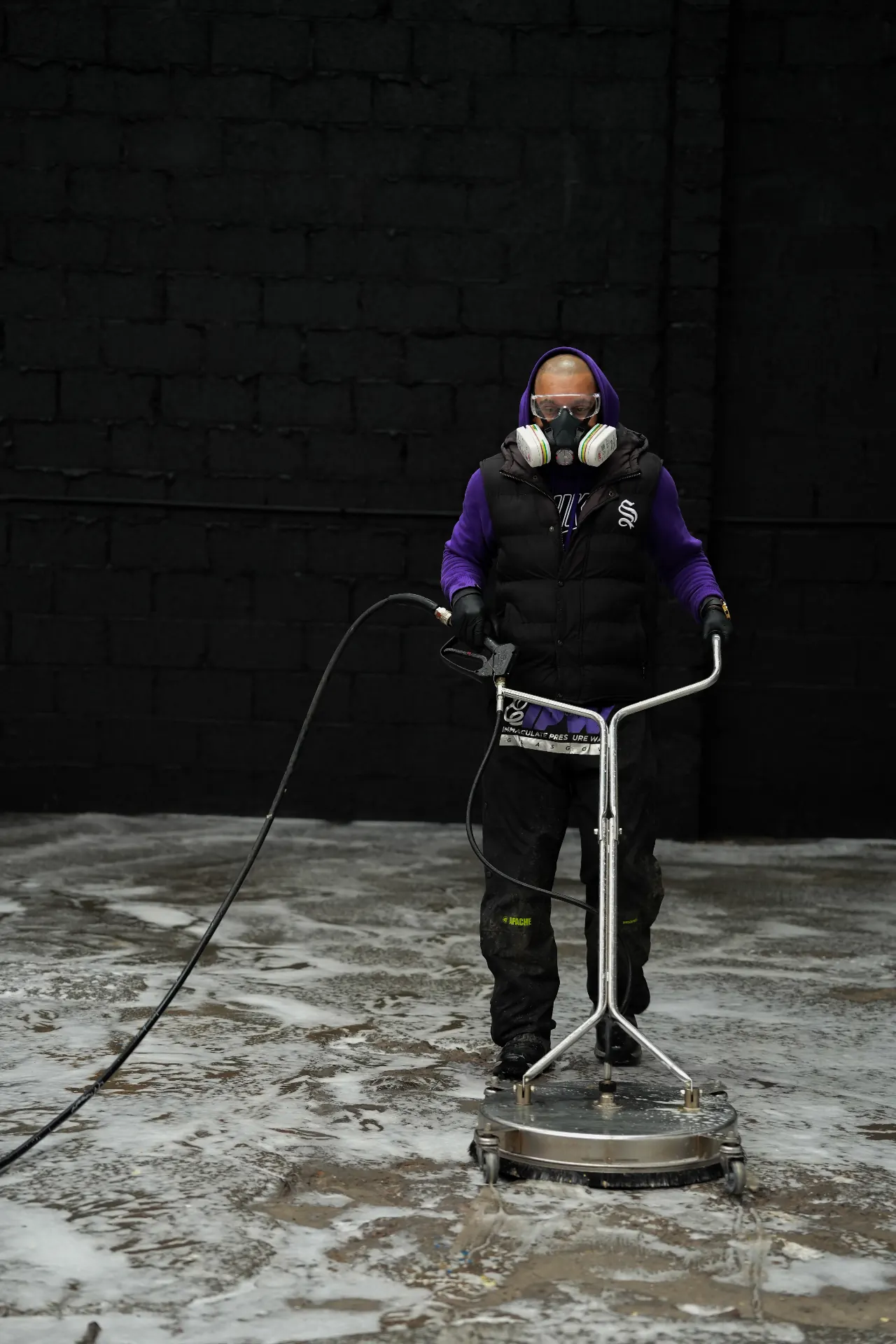 Wide commercial photograph of a worker power washing a floor in a dark black industrial setting.